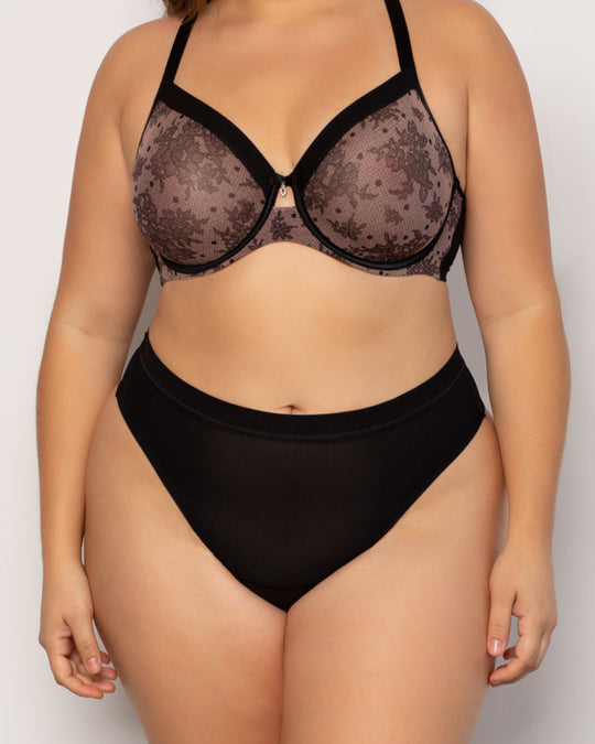 A plus-size model wearing a two-piece lingerie set consisting of a sheer black floral lace underwire bra and solid black high-waisted briefs, photographed against a neutral light gray background.