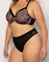 A plus-size model wearing a two-piece lingerie set consisting of a sheer black and brown floral lace bra and solid black high-waisted briefs, photographed against a light gray studio background.