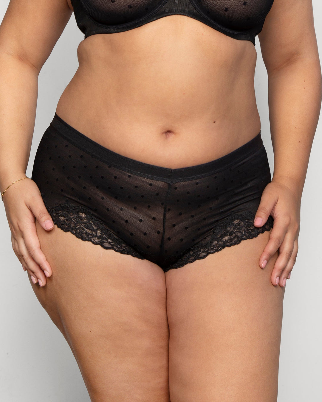 A close-up of a model wearing black sheer mesh panties with small polka dots and a lace leg band, paired with a matching black bra against a light gray background.